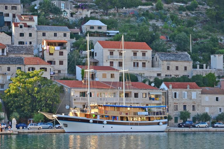 Traditionelles Holzschiff San Snova mit zwei Masten im Hafen in Kroatien, mit Steinhäusern und grünem Hang im Hintergrund.