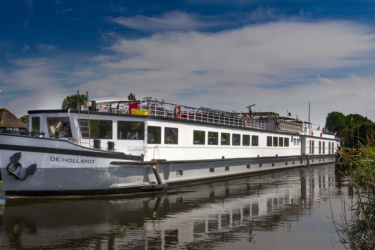 White river cruise ship named De Holland moored on calm water with reeds in foreground and blue sky with white clouds above.