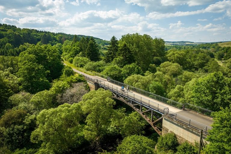 Luftaufnahme einer geschwungenen Betonbrücke am Kyll-Radweg bei Stadtkyll, umgeben von üppigem grünem Wald in der Eifel.