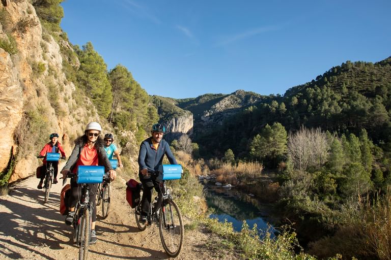 Gruppe von Radfahrern auf einem Flussweg in der Region Valencia, mit Felsklippen und grünen Hügeln unter blauem Himmel.