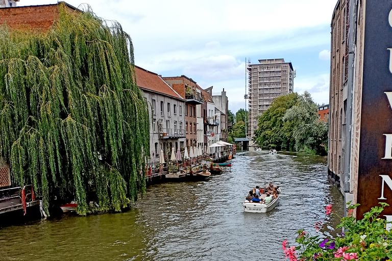 Touristenboot auf Kanal in Brügge mit historischen Gebäuden, Trauerweide und modernem Turm im Hintergrund. Bunte Blumen im Vordergrund sichtbar.