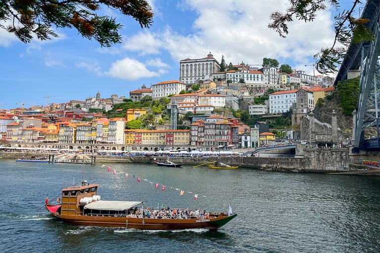 Traditionelles Holzboot mit Passagieren auf dem Douro in Porto, mit bunten historischen Gebäuden am Hang und der Dom Luís I Brücke.