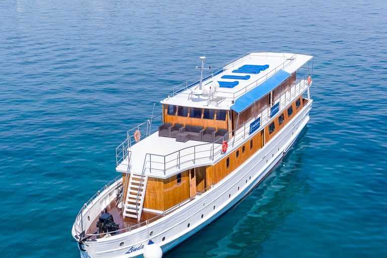 Luftaufnahme der MY Linda, ein weißes Kreuzfahrtschiff mit Holzdeck und blauen Sonnenliegen auf dem Dach, vor Anker in türkisfarbenem Wasser.
