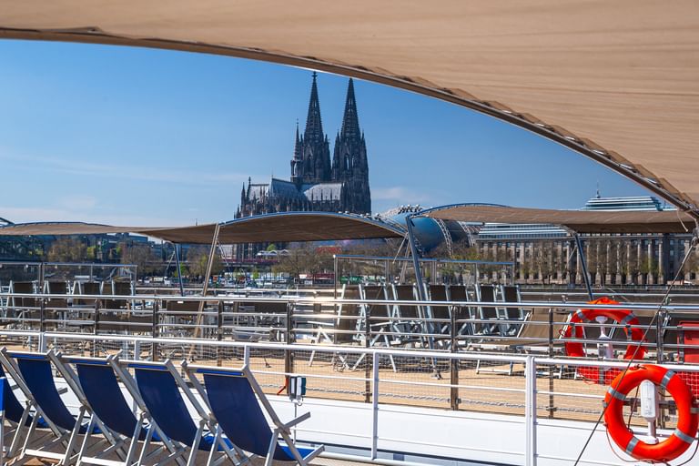 Sun deck of MS Lisabelle with blue deck chairs and red life ring, overlooking Cologne Cathedral's twin spires across the Rhine River.