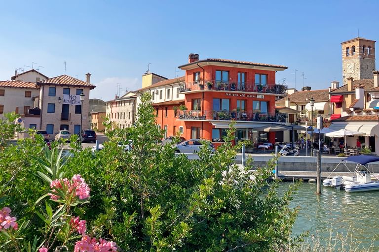 Uferblick auf Marano in Friaul mit bunten Gebäuden, Kirchturm, Kanal mit Booten und rosa blühenden Büschen im Vordergrund.