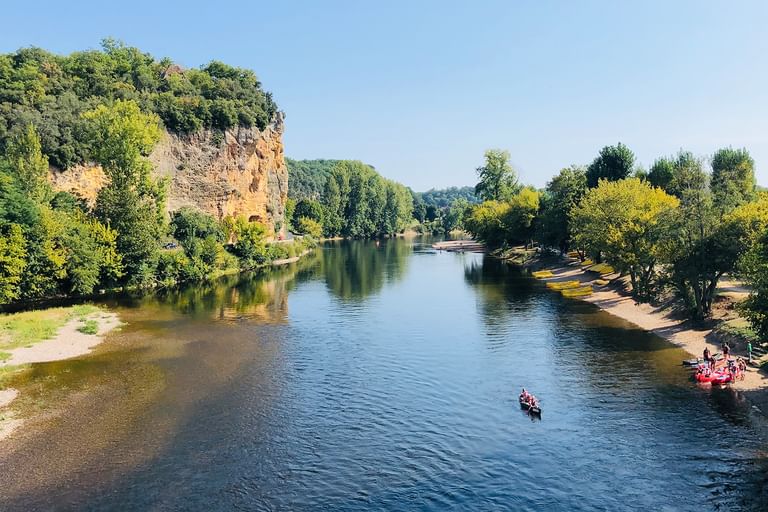 Blick auf die Dordogne mit Kanus auf dem Wasser, Sandstränden an beiden Ufern, üppigen grünen Bäumen und einer Kalksteinklippe links.
