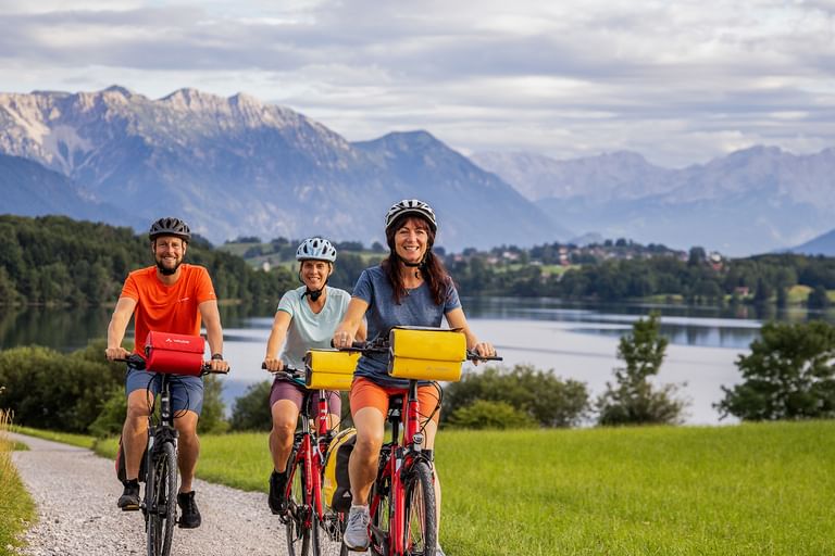 Drei Radfahrer mit Helmen fahren auf einem Weg entlang eines Sees in Österreich. Berge und grüne Wiesen im Hintergrund unter bewölktem Himmel.