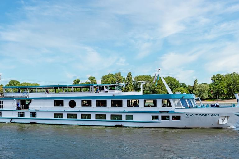 Weißes Flusskreuzfahrtschiff MS Switzerland mit blauem Zierstreifen auf ruhigem Wasser. Bäume und blauer Himmel im Hintergrund.