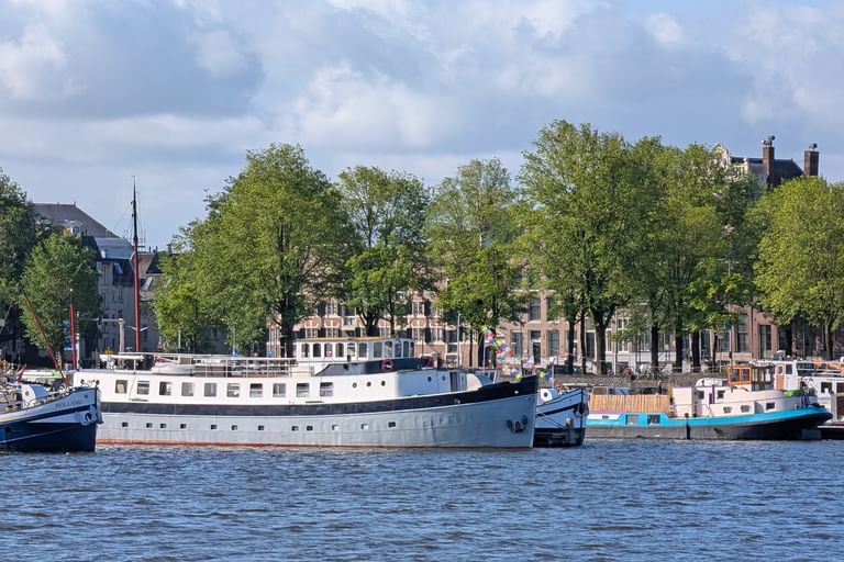 Weiß-blaues Hausboot Floor van Aemstel im Hafen mit Bäumen und historischen Gebäuden am Ufer unter bewölktem Himmel.
