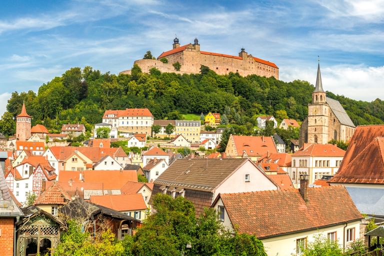 Panoramablick auf Kulmbach mit historischer Burg auf dem Hügel über der Stadt mit roten Ziegeldächern, Kirchturm und grünen Wäldern.
