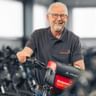 Uwer Hanusch mit Brille und grauem Poloshirt, lächelnd am Fahrradlenker in einer Werkstatt mit vielen Fahrrädern im Hintergrund.