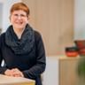 Meike Engling mit kurzen roten Haaren und Brille, trägt ein schwarzes Oberteil und gemusterten Schal, lächelt an einem Schreibtisch in einem hellen Büro.