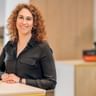 Irene Godoy Marin mit lockigem braunem Haar, in schwarzem Hemd und Uhr, lächelnd an einem weißen Tisch in einem modernen Büro.