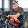 Robert Goldberg wearing glasses and blue cap, leaning on bicycle with red bag in fitness center with exercise bikes in background.