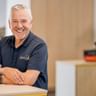 Uwe Klemme mit grauen Haaren trägt ein dunkles Poloshirt und lächelt an einem Tresen. Er trägt eine Smartwatch in einem hellen Büroraum.