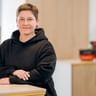 Romy Kellert mit kurzen braunen Haaren in schwarzem Hoodie, stehend an einem hellen Holztisch in einem modernen Büroraum.