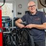 Herms Preuss mit Brille und grauem Poloshirt lächelnd in einer Fahrradwerkstatt, arbeitet an einem Fahrrad mit rotem Montageständer und Werkzeugen.
