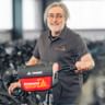 Hans Wüst with glasses and gray hair, wearing a dark polo shirt, standing with a bicycle in a workshop filled with bikes.