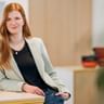 Phyllis Wiechmann with long red hair wearing a beige blazer and black top, standing in a modern office with blurred background.