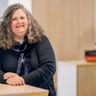 Britta Krefeldt with shoulder-length curly gray hair, wearing a dark blazer and patterned scarf, smiling in a bright office setting.