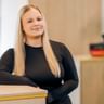 Cassandra Morla Silva mit langen blonden Haaren trägt ein schwarzes Langarmoberteil und steht an einem Empfangstresen in einem hellen Büroraum.
