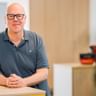 Markus Hüneke mit schwarzer Brille und grauem Poloshirt, sitzt an einem hellen Holztresen in einem hellen Büroraum.