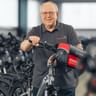 Janos Raduly mit Brille und grauem Poloshirt steht mit einem Fahrrad mit roter Tasche in einer Lagerhalle voller Fahrradreihen.
