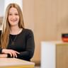 Yasmine Rohlfs mit langen blonden Haaren trägt ein schwarzes Langarmoberteil und eine Halskette, lächelt an einem Holztisch in einem modernen Büro.