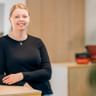 Janina Bronn mit zurückgebundenen blonden Haaren, trägt ein schwarzes Langarmoberteil und eine Halskette, lächelt in einem modernen Büro.
