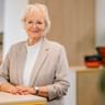 Anke Tammen with short blonde hair and glasses, wearing a beige blazer over a white turtleneck, standing in a bright office.