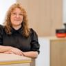Marscha Büsing with curly blonde hair wearing a black blouse, sitting at a light wooden desk in a bright office with blurred background.