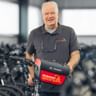 Carsten Rehbein steht mit einem Fahrrad in einer Lagerhalle voller Fahrradreihen. Er trägt ein graues Poloshirt und hält eine rote Tasche.
